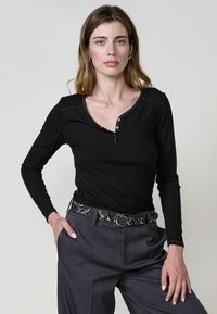 Haut noir à manches longues à côtes avec un col dégagé et des accents de boutons, associé à un pantalon gris ajusté et une ceinture à motifs.