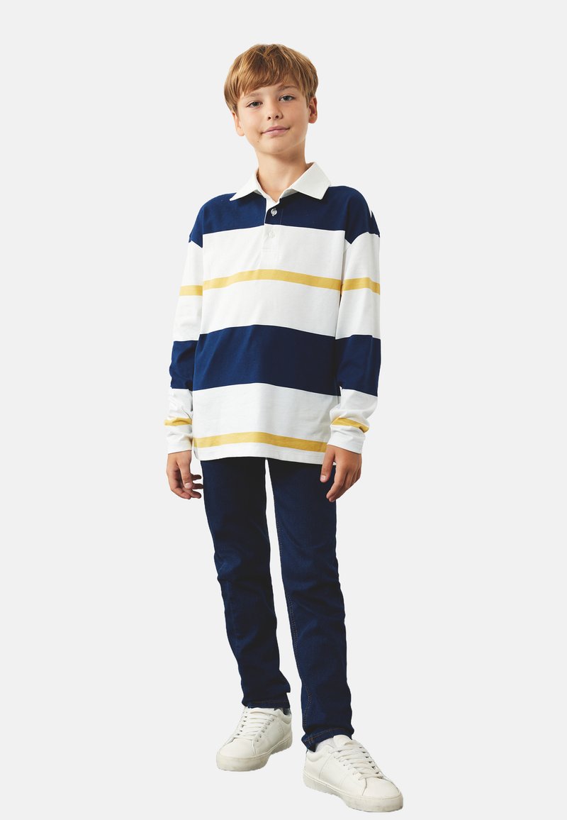 Polo a maniche lunghe a righe bianche e blu navy con una striscia gialla, abbinata a jeans blu scuro e sneakers bianche. Vestibilità casual e texture morbida.