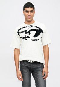 Balta T-krekls ar īsām piedurknēm, uzsverot izteiksmīgu melnu grafisku dizainu uz krūtīm. Valdots ar tumši pelēkām džinsiem un melnu jostu.