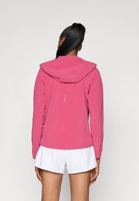 Veste à capuche rose avec une texture lisse et une coupe ajustée, dotée d'accents réfléchissants et d'un design de dos allongé. Associée à un short blanc.