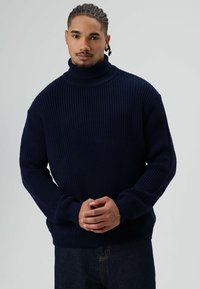 Marineblauwe gebreide coltrui met geribbelde structuur, losse pasvorm en lange mouwen; gecombineerd met donkere spijkerbroek.