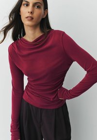 Dlhý rukáv top v hlbokej burgundskej látke, so zberaným výstrihom, priliehavým dizajnom a hladkou textúrou, skombinovaný s tmavými nohavicami.