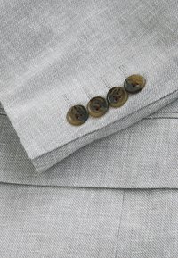 Manche de costume gris clair avec quatre boutons marron foncé marbrés cousus en ligne près du poignet sur un tissu texturé.