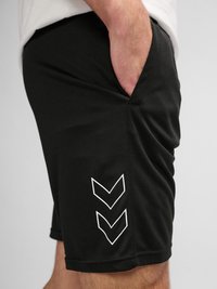 Gros plan d'une personne portant un short noir avec deux logos chevrons blancs sur la jambe gauche, main reposant dans la poche.
