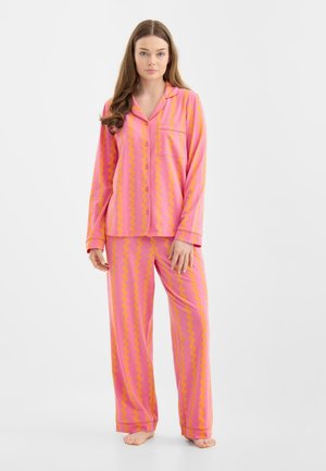 Vrouw staat blootsvoets en draagt een roze met oranje gestreept pyjamapak met knopen, lange mouwen en wijde pijpen tegen een witte achtergrond.