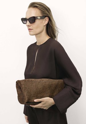 Barna textúrájú, foltos mintázatú clutch táska, egy kézben tartva; sötét, hosszú ujjú felsővel viselve, melyen cipzár található. Fekete napszemüveg.