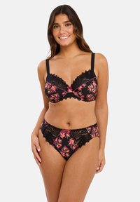 Femme souriante portant un soutien-gorge en dentelle florale noire et un bas assorti devant un fond blanc uni.