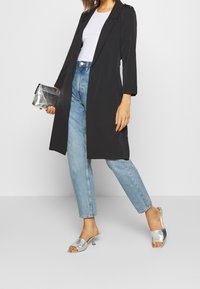 Manteau long noir cintré, escarpins argentés à enfiler, jean taille haute bleu clair, complétés par une pochette argentée.