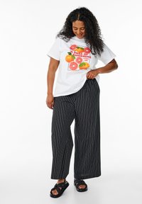 T-shirt bianca con grafica di frutta e testo "Una dose di sole ogni giorno", abbinata a pantaloni larghi a righe verticali nere e sandali neri.