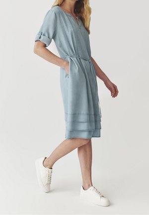 Kobieta ubrana w jasnoniebieską sukienkę za kolano z podwiniętymi rękawami i białe trampki, idąca z jedną ręką w kieszeni.