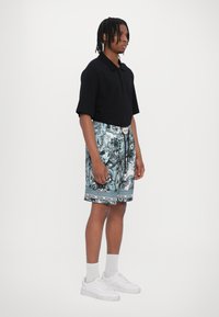 Polo shirt nero, pantaloni corti a fantasia blu e bianca con cordoncino, sneakers bianche e calzini grigi. Design semplice e dal taglio rilassato.