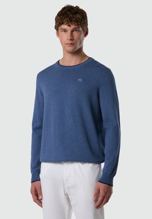 Junger Mann trägt einen blauen Rundhalspullover und weiße Hosen, steht vor einem schlichten hellgrauen Hintergrund.