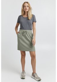 Tricou gri asortat cu o fustă texturată de un verde deschis, având talie cu șnur, prezentat alături de teniși bej și gri.