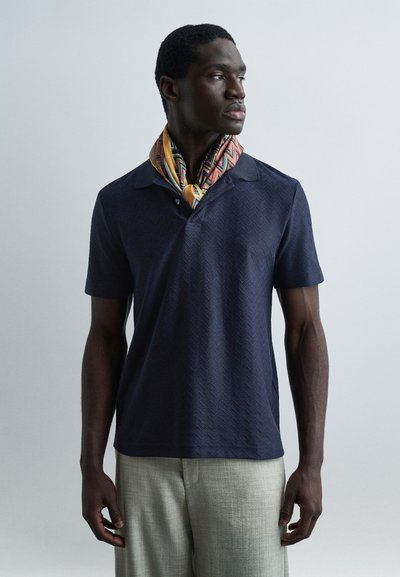 Homme portant un polo texturé à manches courtes bleu marine, pantalon beige clair et écharpe colorée à motifs, regardant vers sa droite sur un fond gris.