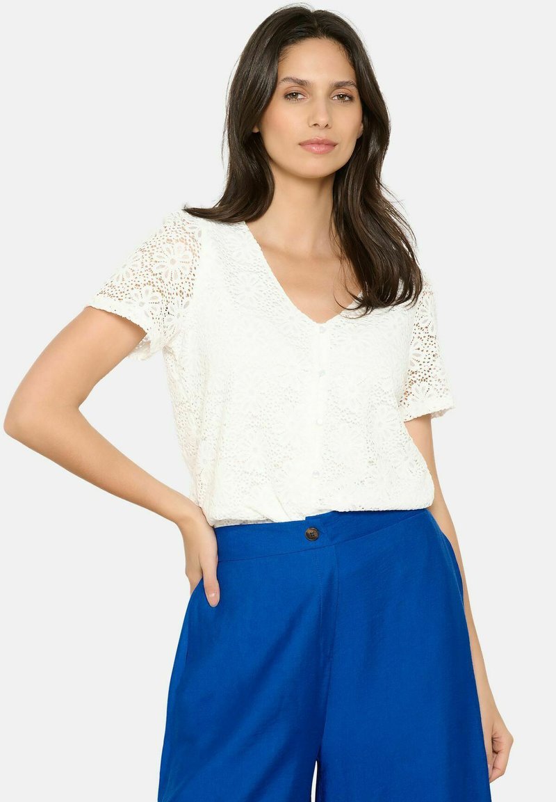 Blusa bianca in pizzo con maniche corte e scollo a V, caratterizzata da dettagli con bottoni sul davanti. Abbinata a pantaloni palazzo blu.