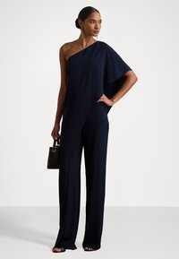 Mørkeblå én-skulder jumpsuit med vide ben, laget av mykt stoff. Inkluderer en liten svart håndveske med et løkkehåndtak.
