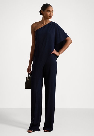 Jumpsuit bleu marine à une épaule avec des jambes larges, fabriqué en tissu doux. Comprend un petit sac à main noir avec une poignée en boucle.