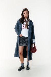 Lungo cappotto grigio a righe su un maglione bianco con testo rosso, gonna nera scintillante, calze grigie, mocassini neri e borsa rossa.