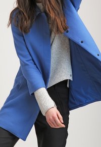 Blue, textured coat with a loose fit and open front. Worn over a light gray knit sweater. Black pants are also visible.