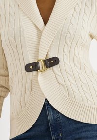 Kreminės spalvos pynimo megztinis su užsegimu – puošniu tamsios odos ir auksinės spalvos sagtimi, dėvimas ant mėlynų džinsų.