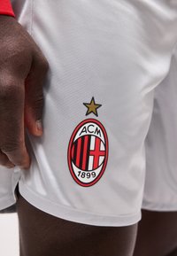 Κοντινό πλάνο ενός σκουρόχρωμου χεριού που ακουμπά σε λευκό σορτς με το έμβλημα του ποδοσφαιρικού συλλόγου AC Μίλαν και ένα χρυσό αστέρι από πάνω.