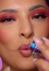 Nahaufnahme eines Gesichts mit rosanem Lidschatten, glänzenden mauvefarbigen Lippen und manikürten Nägeln, die einen mehrfarbigen, schimmernden Lippenstift-Halbstift halten.