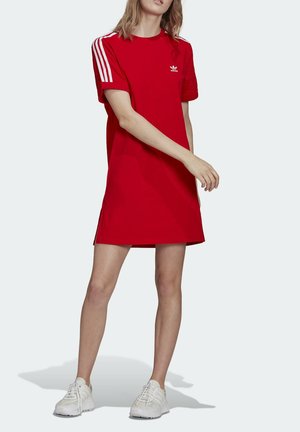 Kobieta w czerwonej sukience-T-shirt marki Adidas z białymi paskami na rękawach, ubrana w białe tenisówki, stoi na jasnym tle.