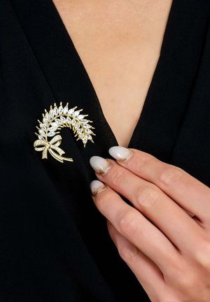 Gold-tone brooch shaped like a leafy plant, featuring clear gemstones and a ribbon detail, attached to a black fabric.