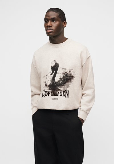 Μαύρος άνδρας που φοράει μπεζ φούτερ με μαύρο γεράκι και το κείμενο "Κοπεγχάγη", συνδυασμένο με μαύρο παντελόνι, στέκεται μπροστά σε μονόχρωμο φόντο.