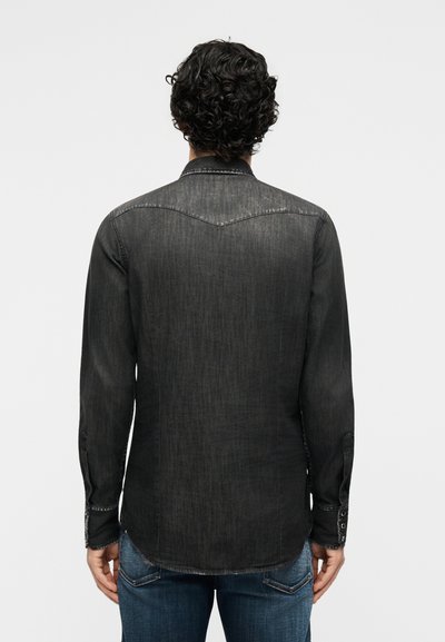 Άνδρας με σγουρά μαλλιά, ντυμένος με ξεθωριασμένο μαύρο τζιν πουκάμισο με μακριά μανίκια και μπλε τζιν, εμφανισμένος από πίσω πάνω σε απλό φόντο.