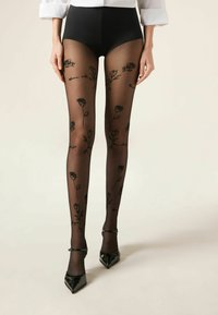 Nogi w przezroczystych czarnych rajstopach z różanymi wzorami i napisami, zestawione z czarnymi butami na obcasie typu Mary Jane oraz czarnymi, wysokim stanem szortami.