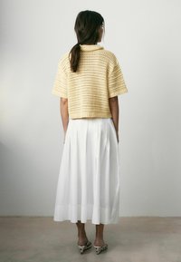 Mujer con cabello oscuro en una coleta baja, vistiendo un top de punto crema de manga corta, falda midi plisada blanca y zapatos de tacón con lunares, de espaldas.