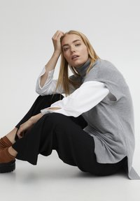 Grijze mouwloze coltrui over een witte lange mouw shirt, gecombineerd met zwarte wijde broek en bruine enkelsokken.