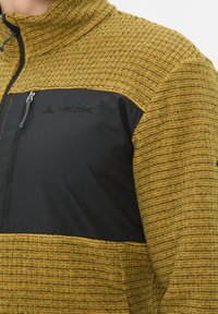 Giacca in fleece gialla con un pannello in poliestere nero. Presenta un colletto alto e una tasca con zip sul lato sinistro del petto. Logo sul pannello.