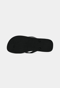 Sandale flip-flop noire vue de dessous avec une semelle texturée et des embouts visibles fixant les lanières, isolée sur fond blanc.