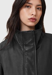Zwarte leren jas met een hoge kraag, gladde textuur en minimale stikseldetails. Gezicht van het model gedeeltelijk zichtbaar, met de nadruk op lippen en ogen.