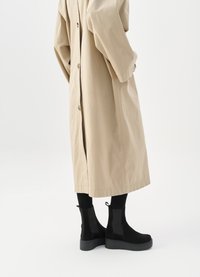 Černé semišové kotníkové boty s elastickými bočními panely a silnými gumovými podrážkami, doplněné béžovým trenčkotem do kolen.