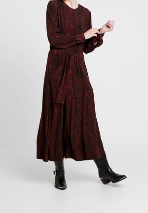Femme portant une robe longue à manches longues, longueur cheville, de couleur rouge foncé avec un motif, une ceinture à la taille, et des bottes noires mi-mollet à bout pointu avec des boucles.
