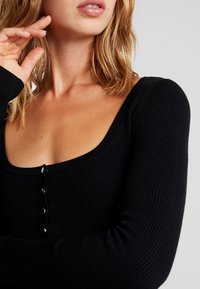 Gros plan d'une femme portant un haut noir à manches longues côtelé avec des boutons, touchant son visage avec la main gauche près de son oreille.