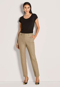 Vrouw in een zwart kort mouwtop, beige geblokte broek, zwarte hakken met gouden punten en grote gouden hoepeloorbellen, die staat met één hand in haar zak.