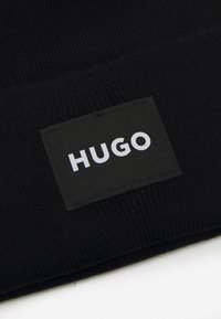 Μαύρο πλεκτό σκουφάκι με ορθογώνια μαύρη υφασμάτινη ετικέτα με το λευκό κείμενο "HUGO" κεντραρισμένο πάνω της. Ψηλή απαλότητα, άνετη εφαρμογή.