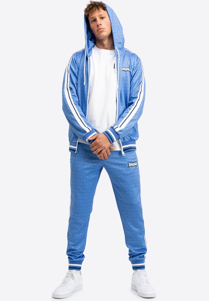 Blauer Trainingsanzug mit geometrischem Muster, bestehend aus einer Kapuzenjacke und Jogginghosen mit Kordelzug. Weiße Streifen entlang der Ärmel und den Seiten der Hose.