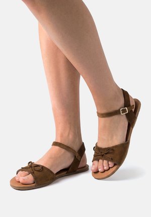 Sandalias planas de ante marrón con tiras al tobillo y detalle de lazo frontal, usadas en pies cruzados sobre un fondo neutro.