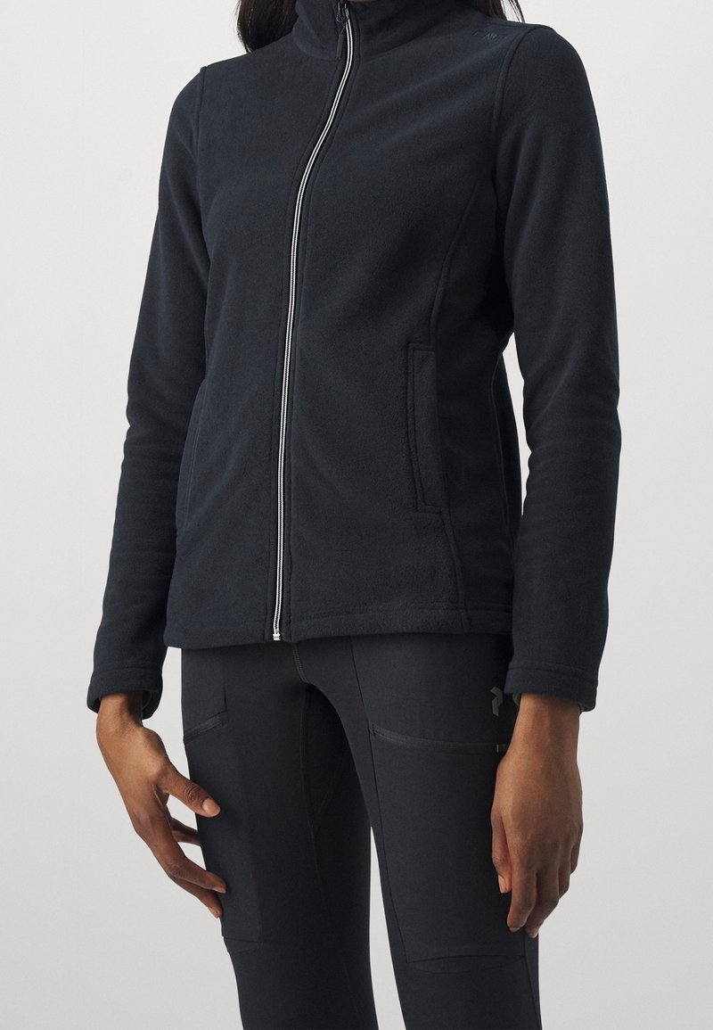 Personne portant une veste polaire noire zippée et un pantalon noir ajusté avec des poches latérales, debout devant un fond clair uni.