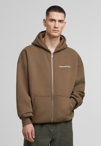 Brun zip-up hættetrøje med snoretræk i hætten, frontlommer og ordet "PROHIBITED" trykt i hvidt. Blødt stof, afslappet pasform.