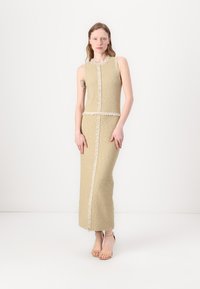 Knelange, ermeløs kjole i strukturert beige stoff, med hvit frynsebesetning langs sømmene og en tettsittende silhuett.