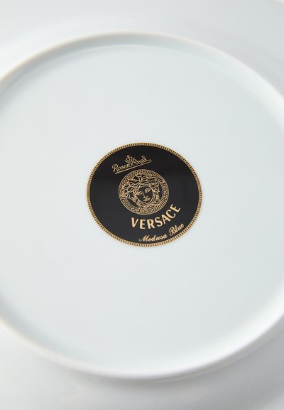 Balta porcelāna šķīvis ar melnu apaļu logotipu centrā ar zelta akcentiem. Logotipā attēloti vārdi "Versace" un "Medusa Blue".