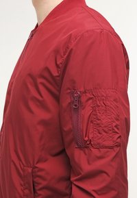 Rote Bomberjacke mit einer Reißverschlusstasche am linken Ärmel, geripptem Kragen und glatter, glänzender Textur.