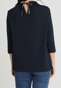 Top azul marino con escote fruncido y detalle de lazo en la parte trasera. Presenta mangas tres cuartos y un corte holgado. Textura suave, diseño casual.