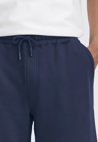 Temno modre trenirka hlače iz mehke tkanine z elastičnim pasom in vrvico za zategovanje. Vključujejo stranske žepe in gladek, teksturiran zaključek.